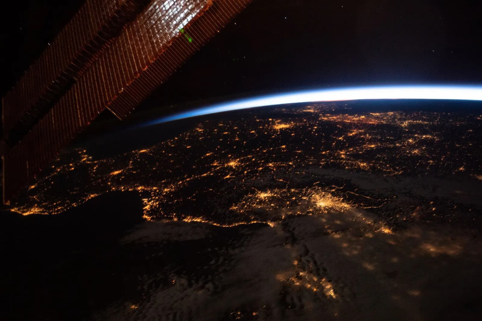 Western Europe at night seen from the International Space Station: orange city lights along the coasts of Spain, France and Italy, a thin blue line of the horizon and a solar panel of the station at the left