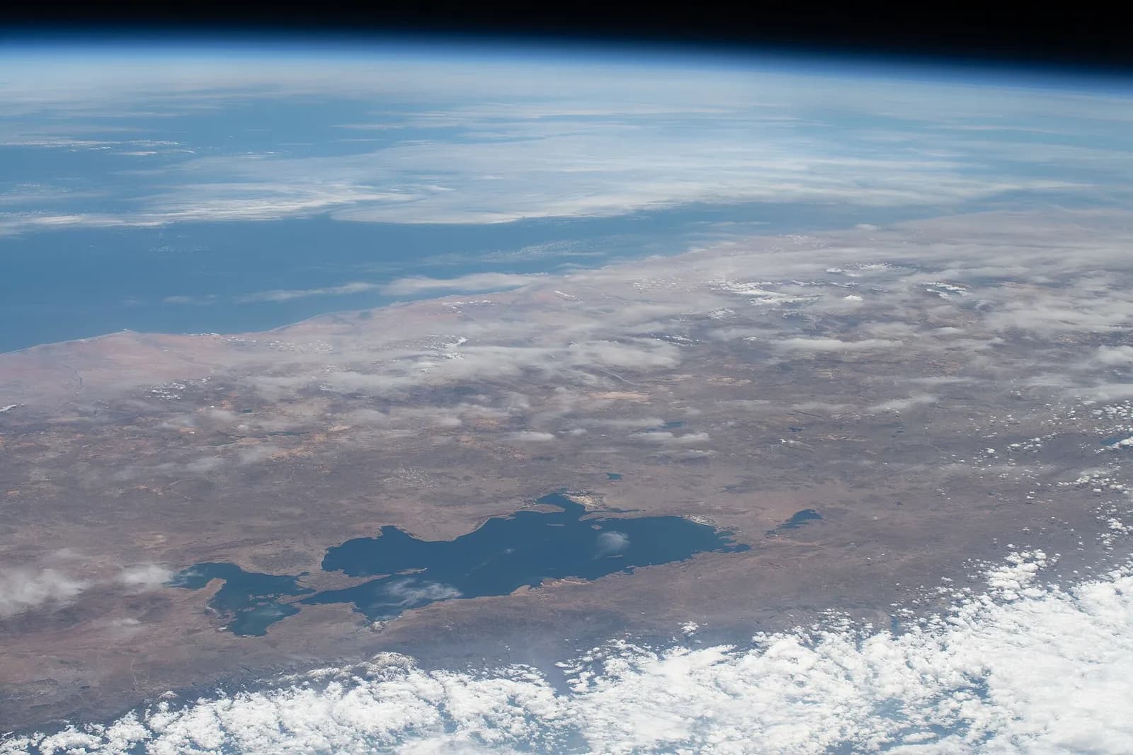 Lake Titicaca between Peru and Bolivia seen from the International Space Station: a dark blue lake on the brown high plateau of the Andes, white clouds in the foreground and the ocean and the curve of the Earth towards the horizon