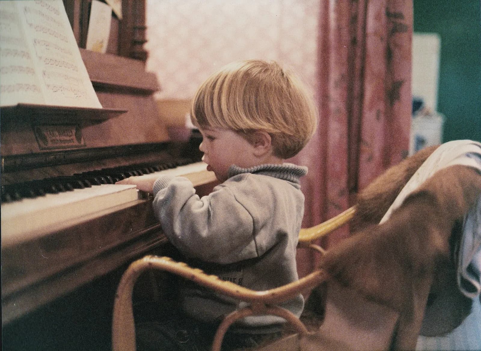 A small child in a grey jumper presses the keys of an old upright piano, with sheet music on the stand