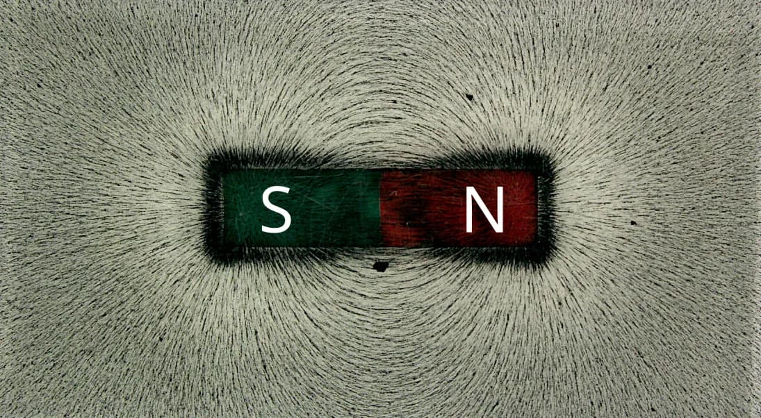 A bar magnet with a green south end and a red north end, marked S and N, surrounded by iron filings that trace curved field lines from one pole to the other