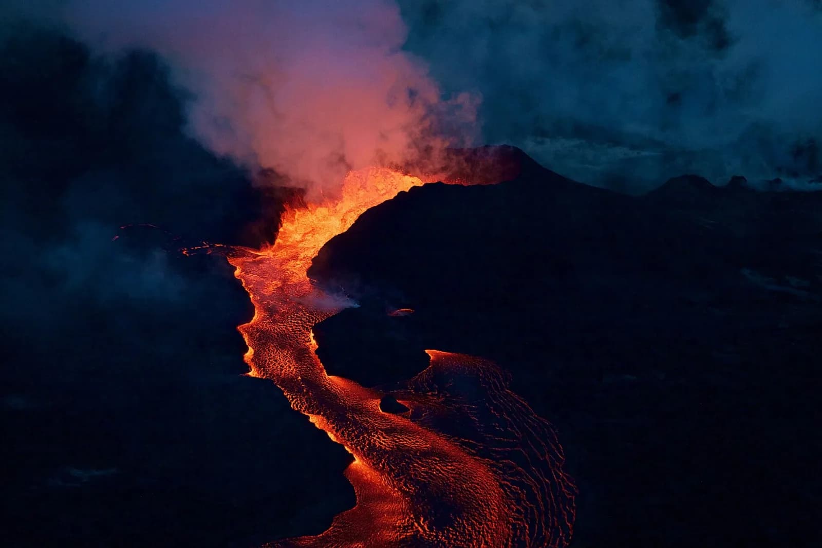 Aerial view at dawn of the Fissure 8 cone on Kīlauea, Hawaii, on 28 June 2018: a glowing river of lava runs from the vent past the dark cone under drifting steam and smoke