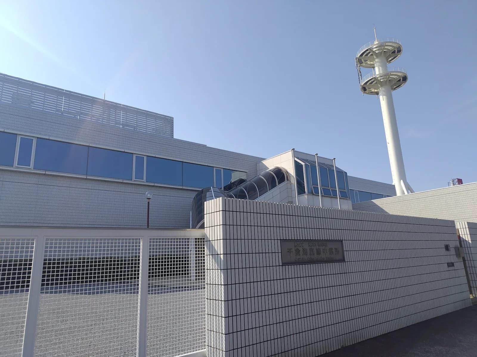 The KDDI Chikura cable landing station in Japan: a white-tiled building with blue windows and a tall white tower, where undersea cables come ashore, under a clear blue sky