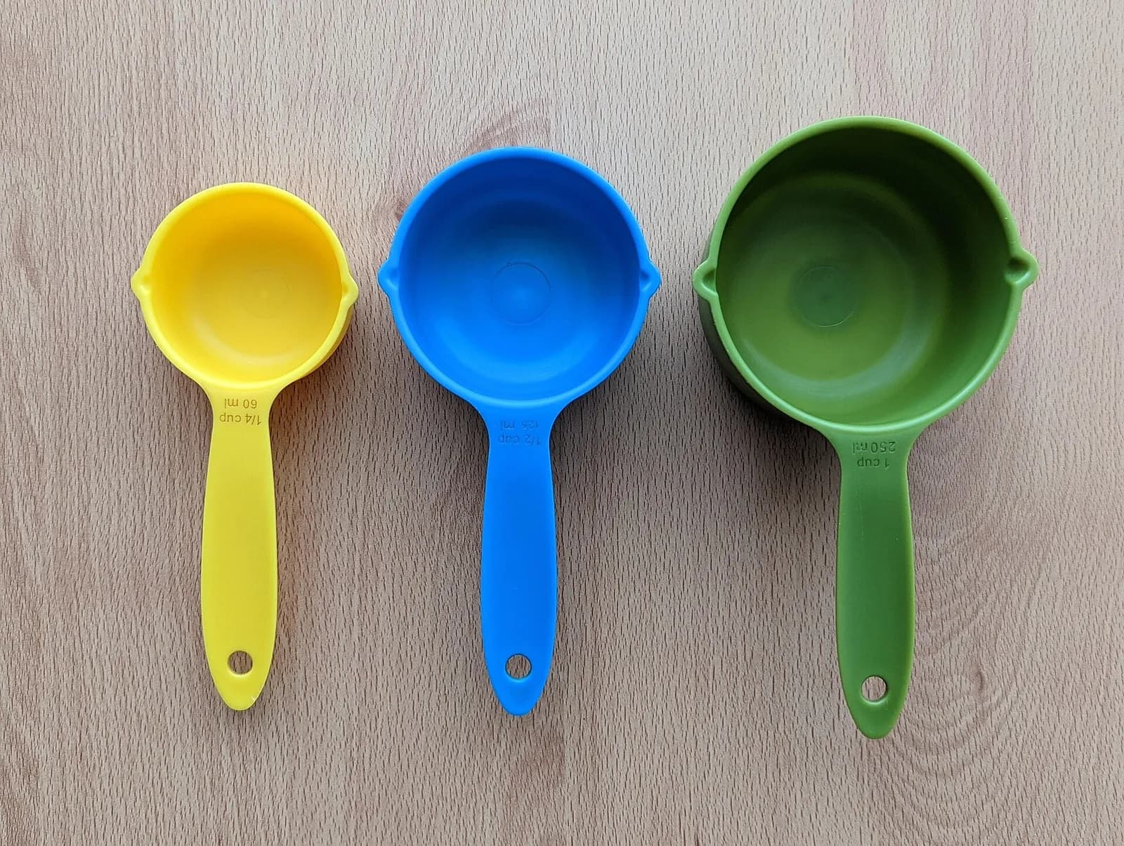 Three plastic measuring cups in a row on a light wooden surface: a yellow 1/4 cup, a blue 1/2 cup and a green 1 cup, each with its size moulded on the handle