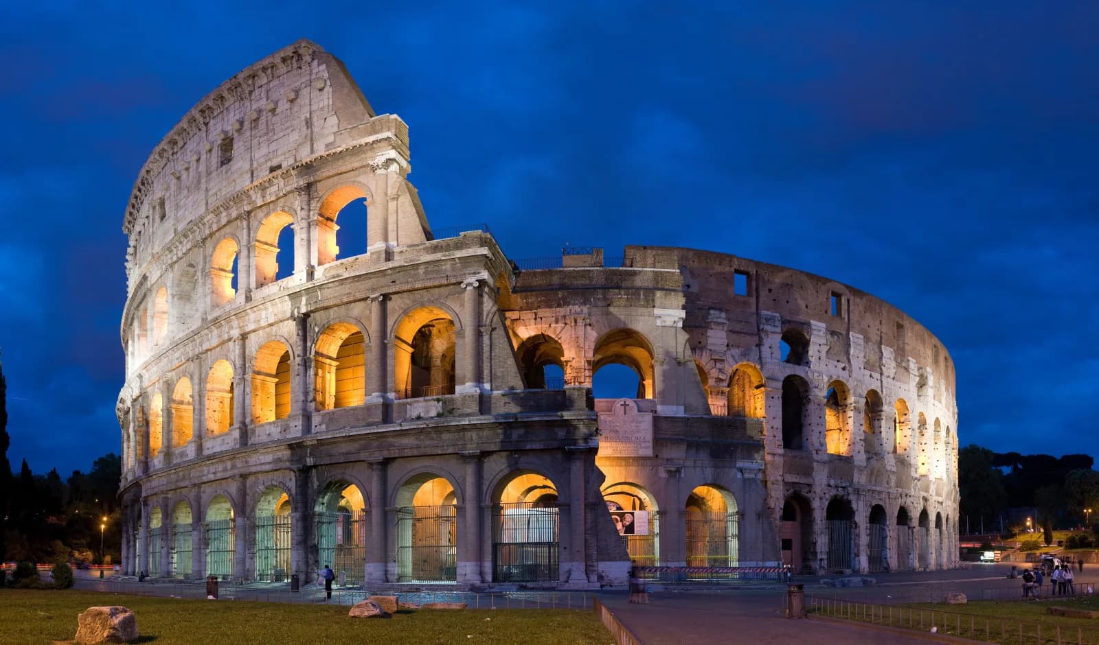The Colosseum in Rome at dusk, its arched outer wall lit warm orange against a deep blue sky