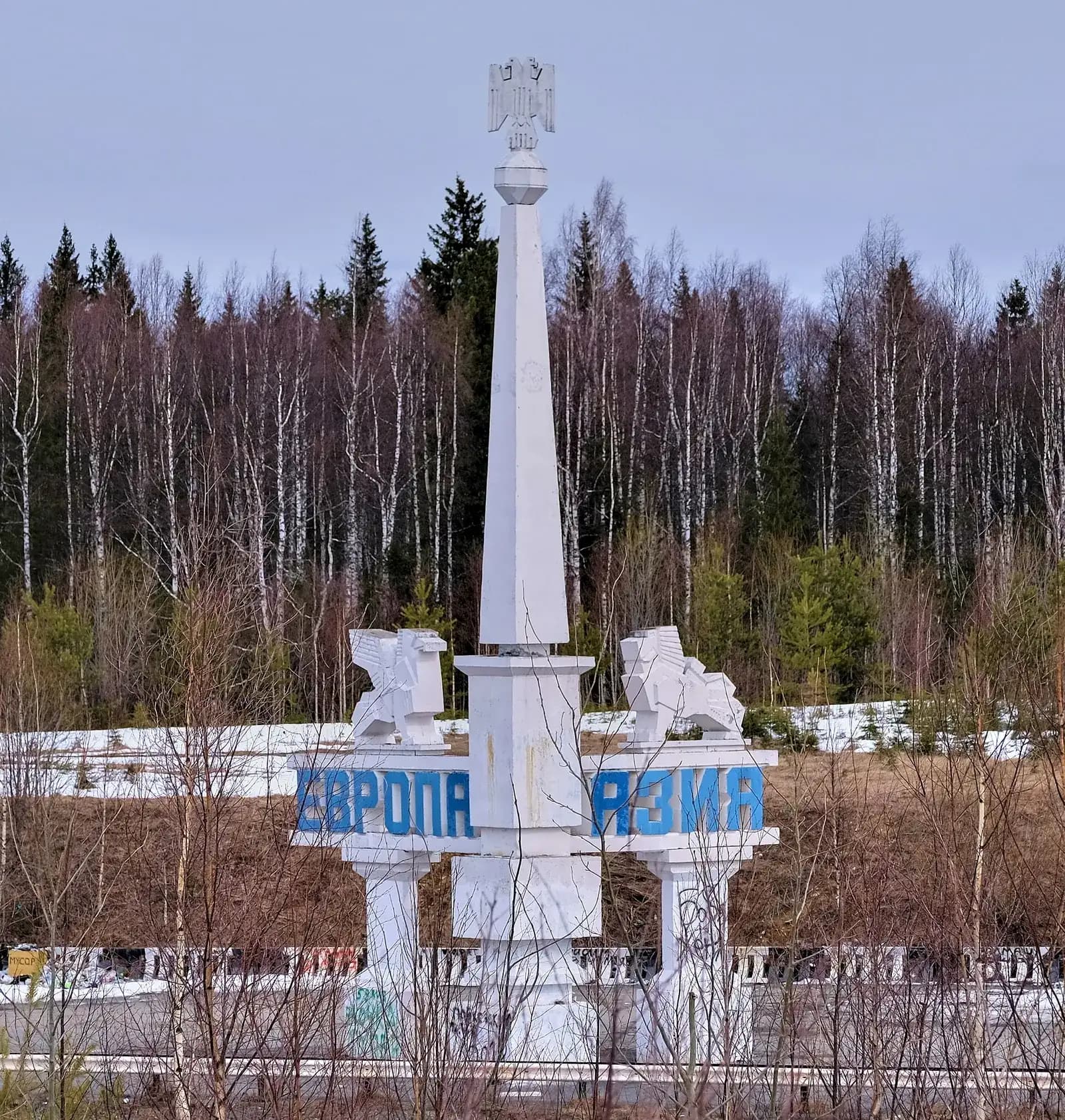 A white obelisk on the Europe–Asia border in the Urals, Russia, with the words ЕВРОПА and АЗИЯ in blue letters, in front of a birch and spruce forest with patches of snow