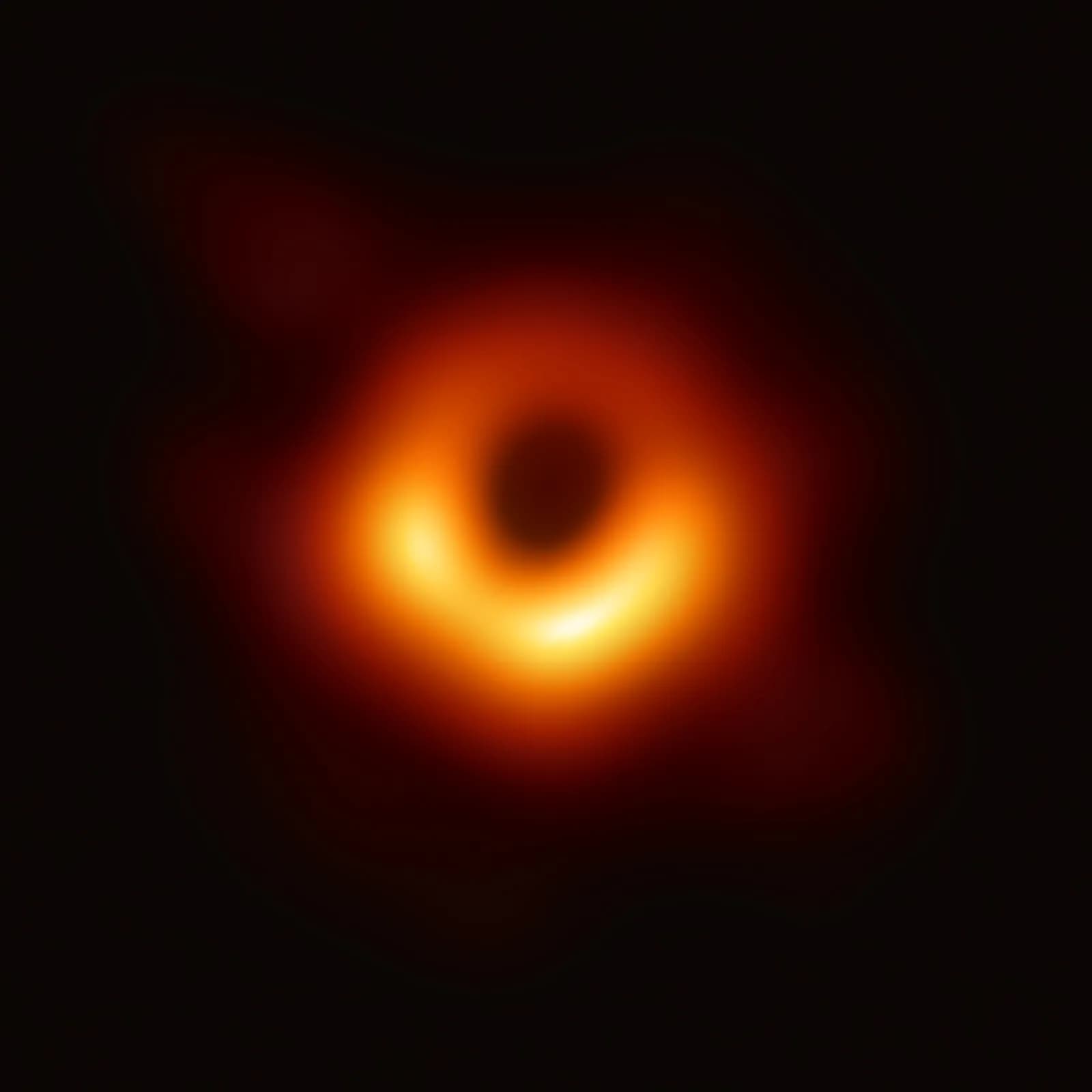 The first image of a black hole, M87*: a lopsided glowing ring of orange and yellow light around a dark central shadow, on black