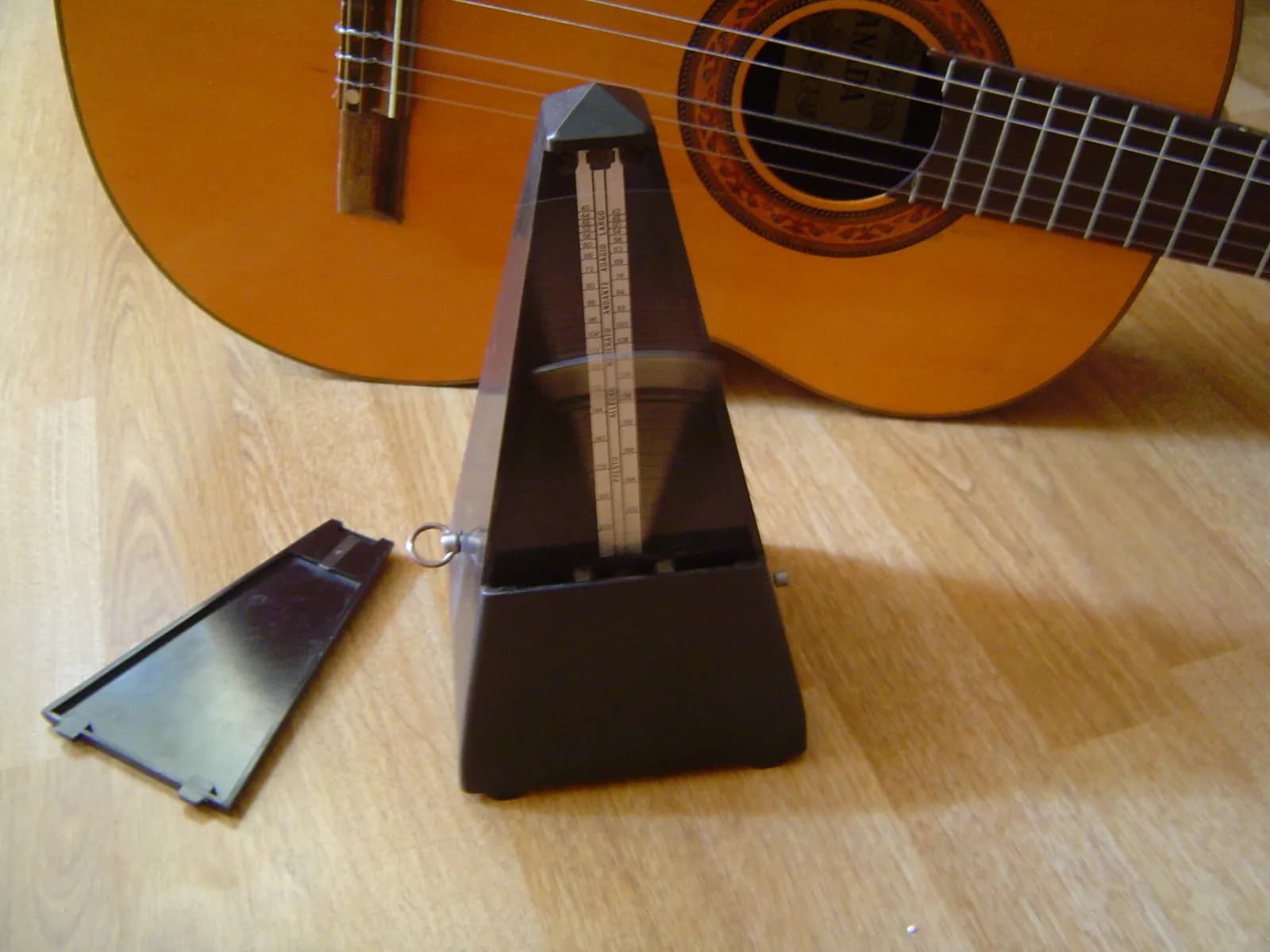 Un metrónomo mecánico de pirámide sin la tapa frontal, con el péndulo en movimiento, delante de una guitarra clásica sobre un suelo de madera