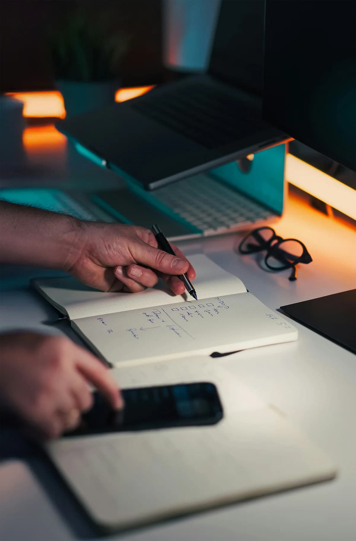 A person making notes beside a laptop and phone