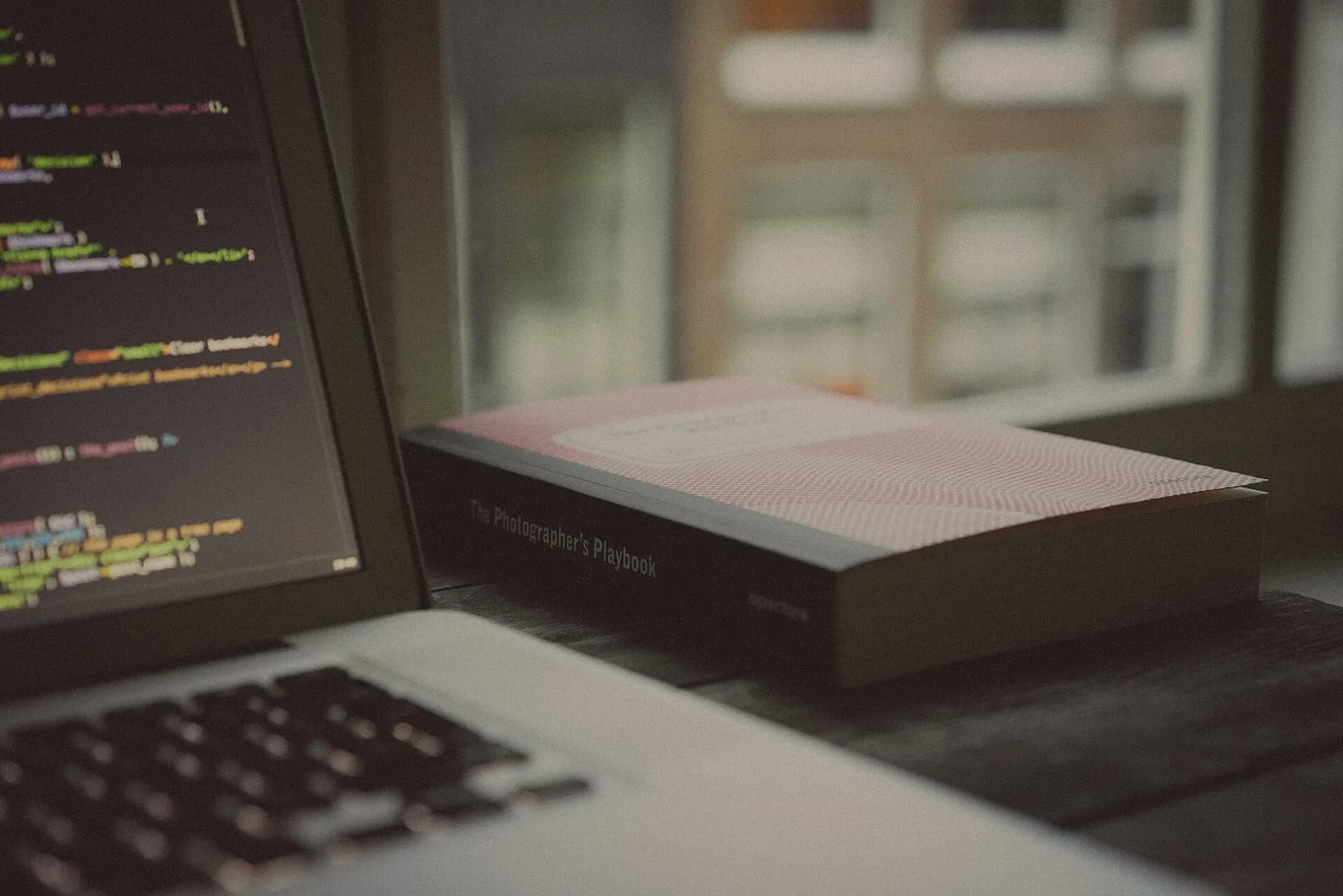 A laptop with code on the screen beside a photography book, on a desk by a window