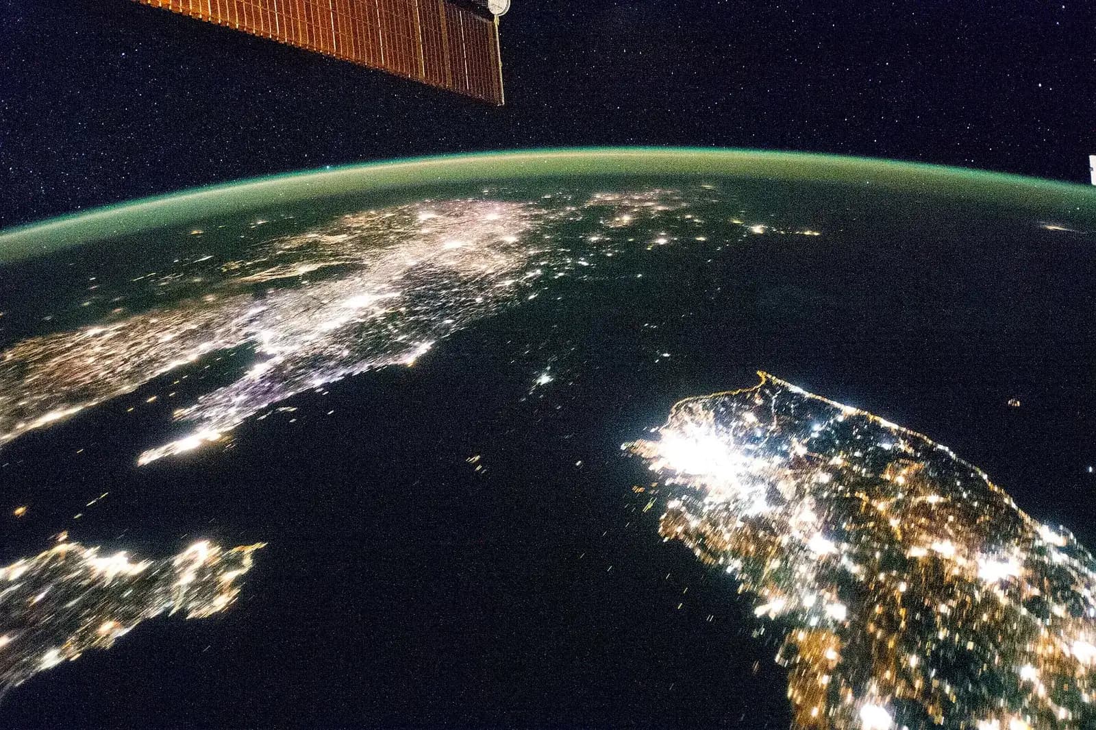 The Korean Peninsula at night from the International Space Station: South Korea glows with city lights at the lower right, North Korea above it is almost dark apart from a small patch of light, and the lights of China fill the left side under a green band of airglow