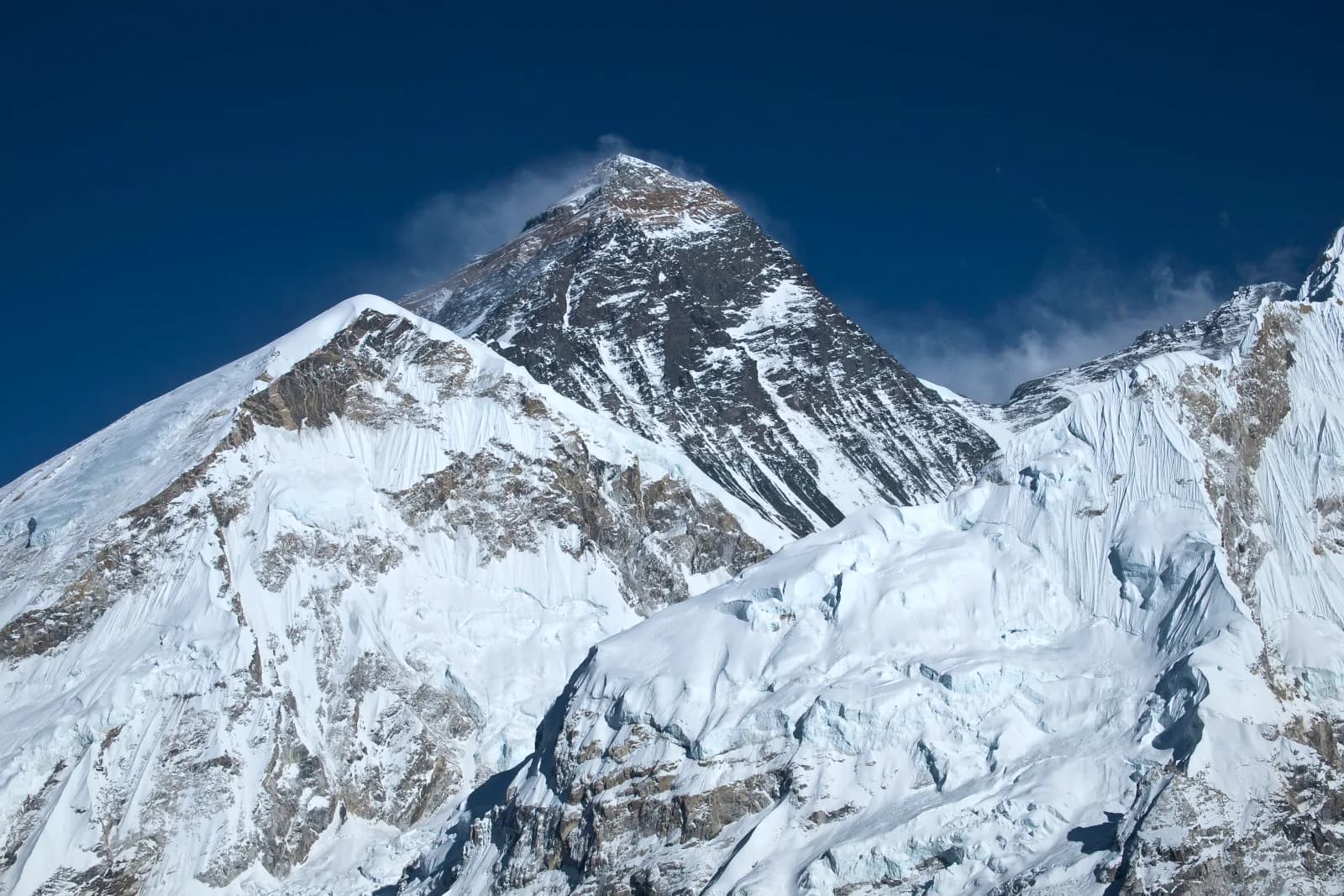 The dark pyramid of the summit of Mount Everest with a plume of wind-blown snow, rising above snow-covered ridges of Lhotse and Nuptse under a deep blue sky