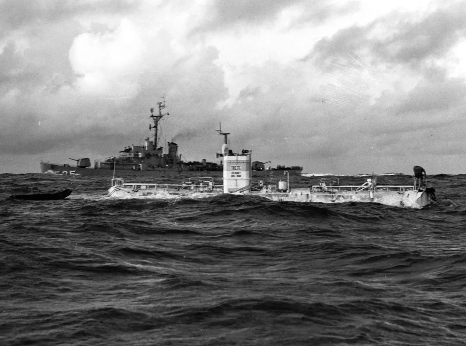 Black-and-white photograph of the US Navy bathyscaphe Trieste floating in rough sea just before its record dive of 23 January 1960, with a crewman on the deck and the destroyer escort USS Lewis in the background under heavy clouds