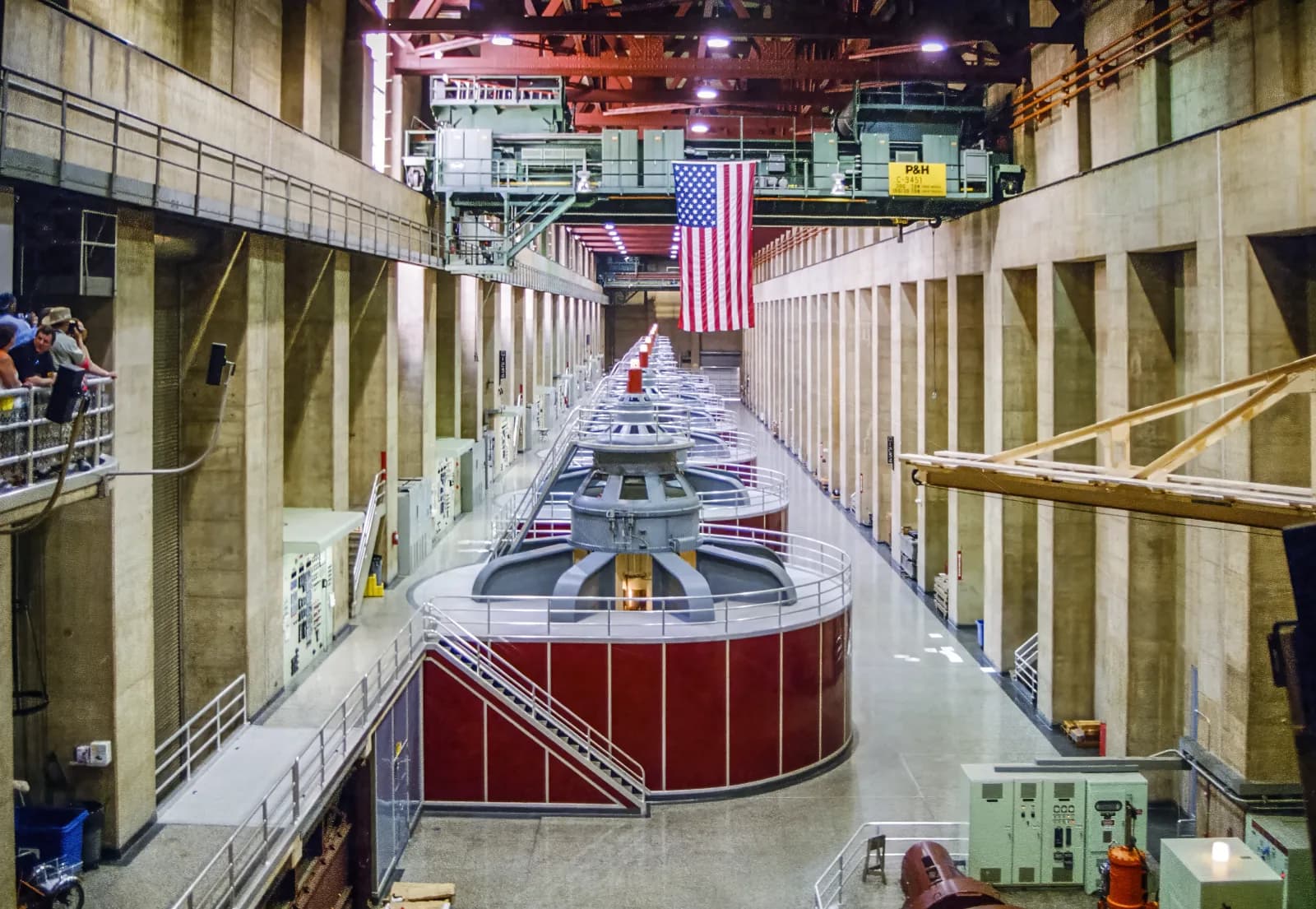 A long concrete hall at Hoover Dam with a row of huge red and grey generator housings, a US flag hanging from an overhead crane and visitors on a balcony at the left