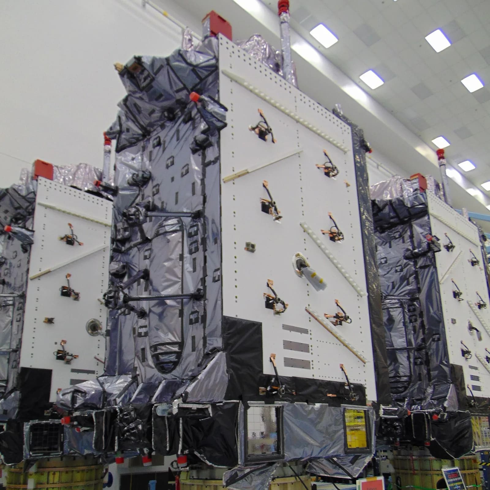 Three GPS III navigation satellites standing side by side in a clean room, boxy white bodies wrapped in silver and black thermal blankets under bright ceiling lights