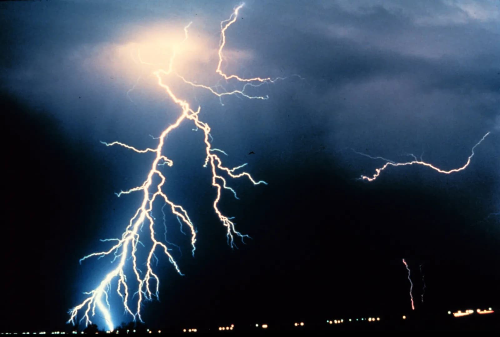 A branching bolt of cloud-to-ground lightning and several smaller strokes lighting dark storm clouds at night over a flat plain with a few distant lights