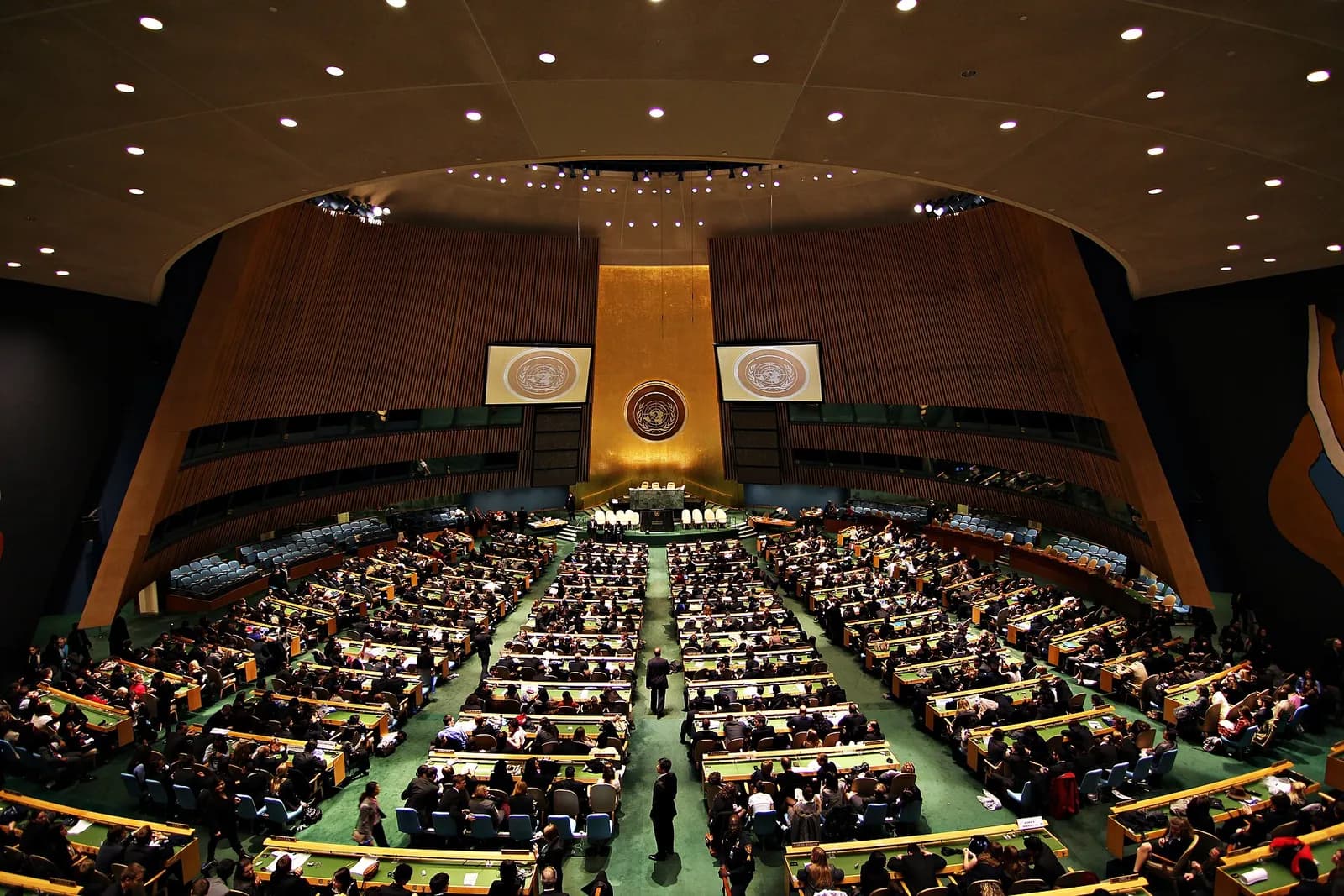 The United Nations General Assembly Hall in New York during a session: rows of delegates at green desks facing the golden wall with the UN emblem