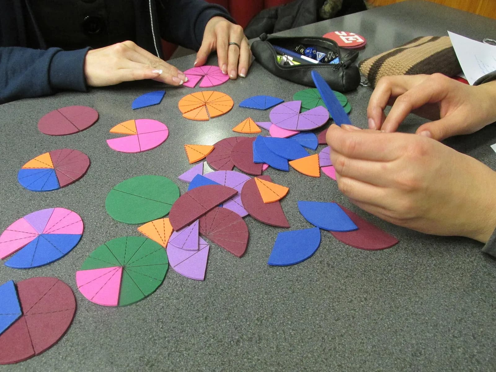 Fraction circles cut from coloured foam paper on a grey table: whole discs, halves, quarters and thinner slices in pink, orange, blue, green and purple, with two people's hands arranging the pieces