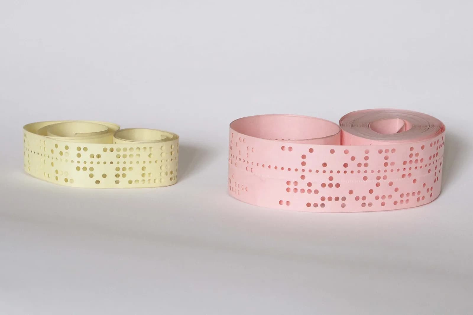 Two rolls of punched paper tape, a narrow pale yellow one and a wider pink one, with rows of holes on a white background