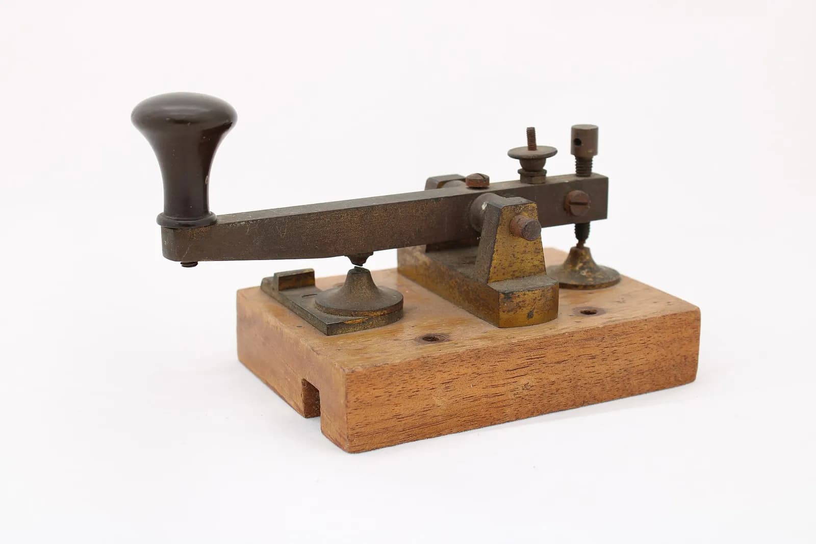 Un antiguo manipulador telegráfico con pomo negro y palanca de metal gastada, montado sobre un bloque de madera, con fondo blanco