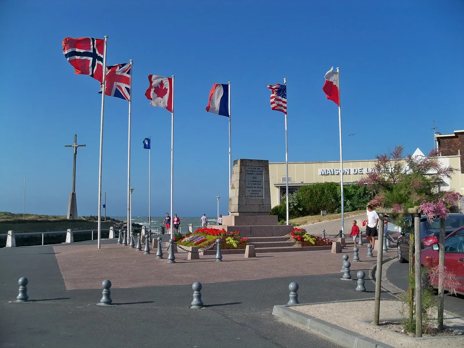 The flags of Norway, the United Kingdom, Canada, France, the United States and Poland flying on tall poles under a clear blue sky around the memorial at Courseulles-sur-Mer in Normandy