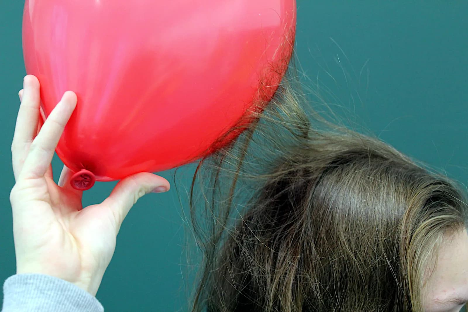 Una mano sostiene un globo rojo ante una pared turquesa mientras mechones de pelo largo se elevan desde la cabeza hacia el globo