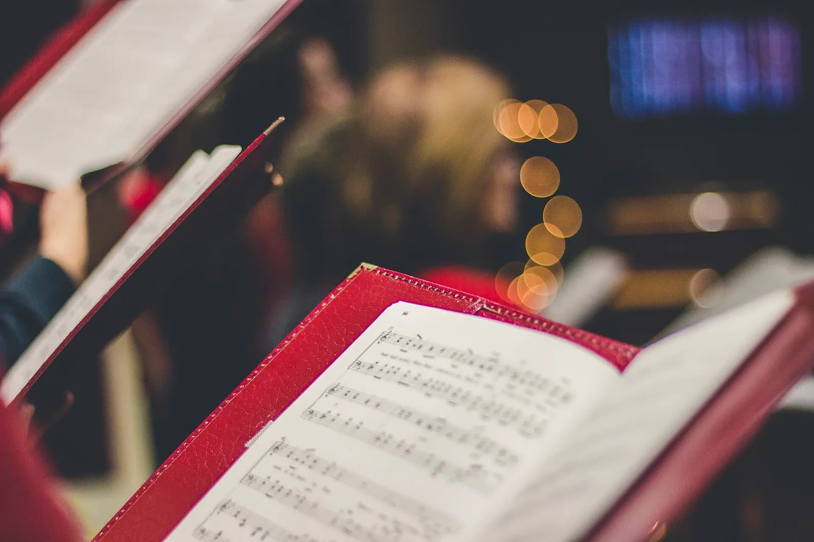 An open choir score in a red folder in the foreground, with blurred singers and warm lights behind