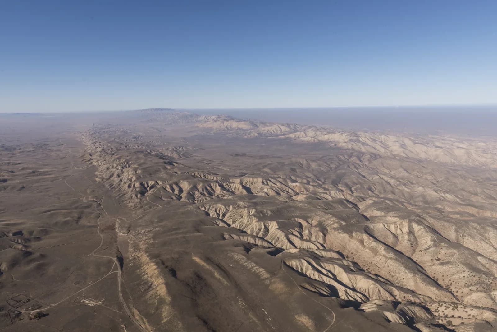Aerial photograph of the San Andreas Fault in California: a long, almost straight line of ridges and dry gullies running into the haze across a pale brown plain under a clear blue sky