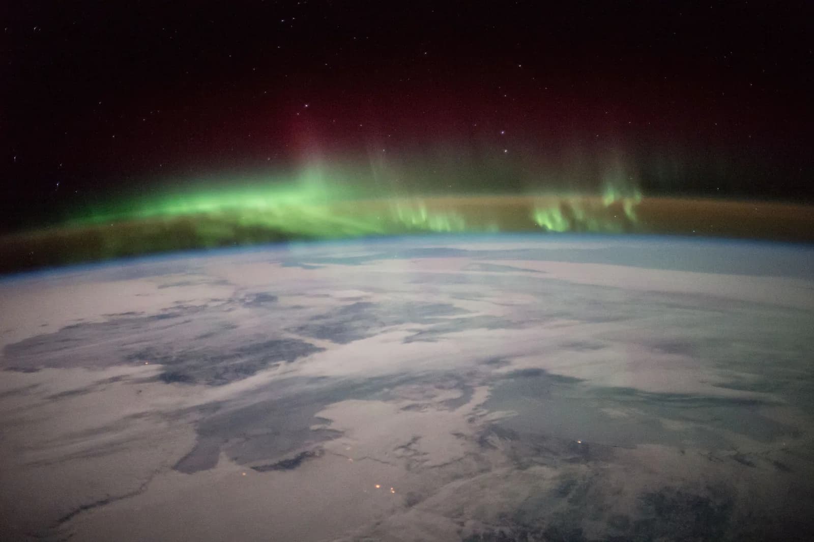 The northern lights seen from the International Space Station over Canada: a green band of aurora with a dark red glow above it, along the edge of snowy land and cloud under the stars