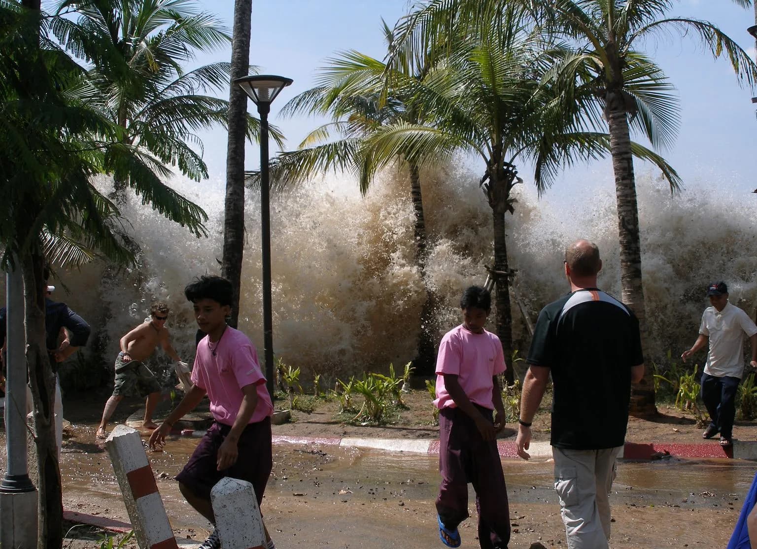 Un muro de agua marrón y espumosa se alza tras las palmeras cuando el tsunami de 2004 llega a Ao Nang, en Tailandia, con personas en primer plano que caminan y corren por una calle inundada
